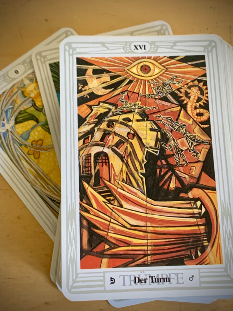 Shows a card deck with the illustrated card 'The Tower' on top. The card consists of an abstract drawing with bright flares, a collapsing building, consuming fire and an all seeing eye above. Behind the deck the card of the fool peeks out.